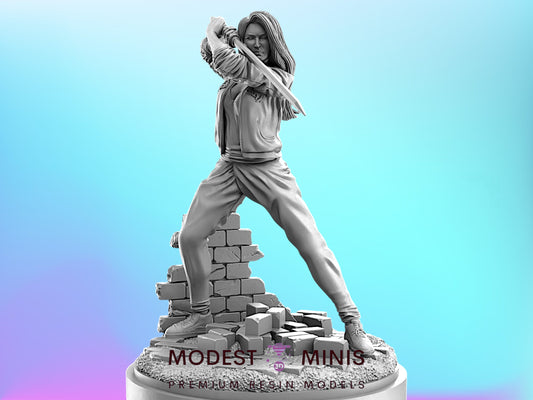 A black and white statue of a woman in a fighting stance, holding a weapon, standing on a base with a textured surface.